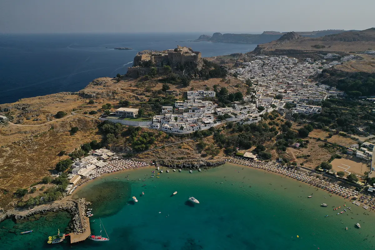 Aerial view of a hillside town with white buildings beside a crescent beach, with a fortress on the ridge above the sea.